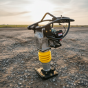 A heavy-duty diesel-powered vibratory plate compactor with a yellow spring and silver engine housing, standing on a gravel surface in an outdoor construction environment at sunset.