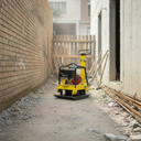 A yellow and black plate compactor machine with a red engine, positioned on a gravel path between a brick wall on the left and a concrete building on the right.