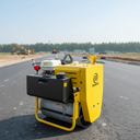 A yellow and black road roller machine photographed outdoors on a freshly paved road, with construction equipment and trees visible in the background.