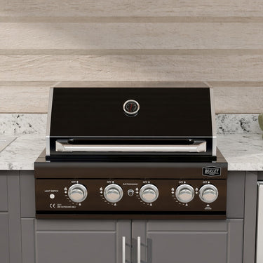 A built-in outdoor gas grill with a black lid and dark brown control panel, installed in a modern kitchen setting with gray cabinets, a white marble countertop, and a light wood backsplash.