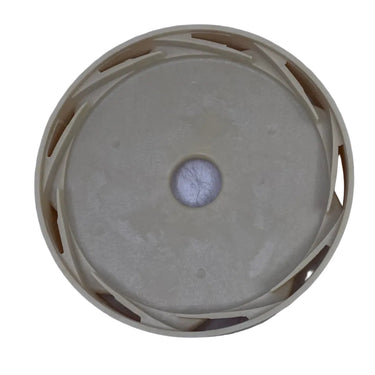 A circular beige plastic cap or cover with a central hole, photographed from above on a scratched light gray surface.