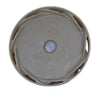 A circular beige plastic cap or cover with a central hole, photographed from above on a scratched light gray surface.