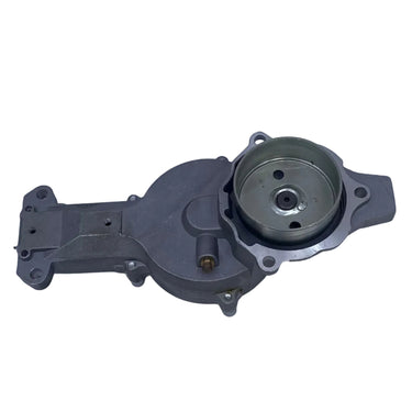 A used metal automotive water pump housing with a pulley and belt cover, photographed on a scratched light gray surface.