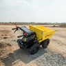 A yellow and black mini dump truck with a rear hopper, photographed outdoors on a dirt construction site under a pale sky. A small worker is visible in the distant background.