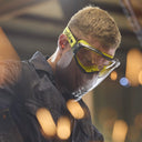 A close-up lifestyle shot of a man wearing yellow and black safety goggles with a clear face shield, set against a warm, blurred industrial background.