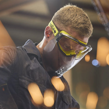 A close-up lifestyle shot of a man wearing yellow and black safety goggles with a clear face shield, set against a warm, blurred industrial background.